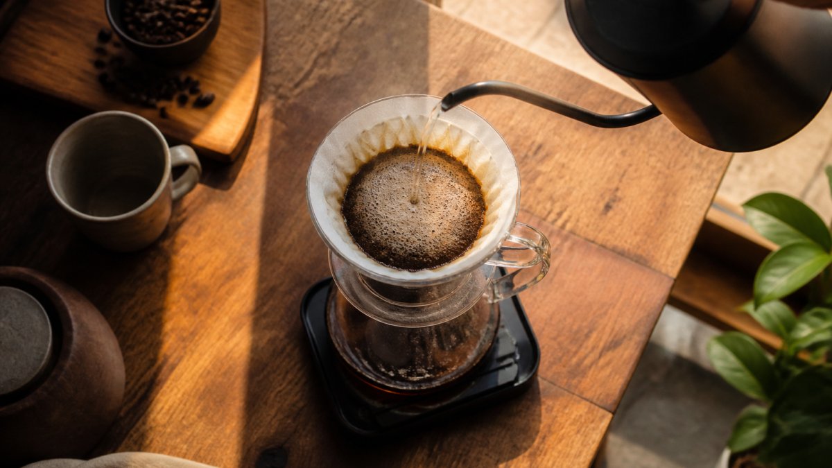Close-up overhead shot of a gooseneck kettle pouring a slow spiral of water into a pour-over dripper on a kitchen scale, steam rising, warm morning light on a wooden counter