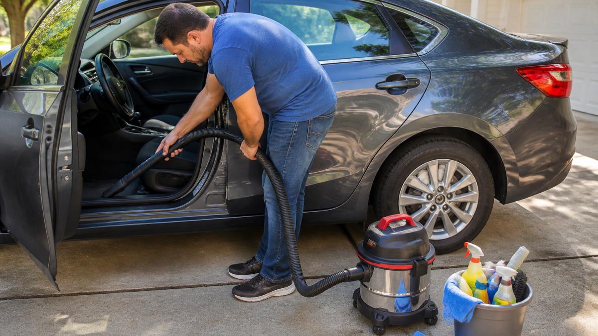 A person vacuuming the interior of a clean used sedan in a driveway, bucket and detailing supplies nearby, bright daylight