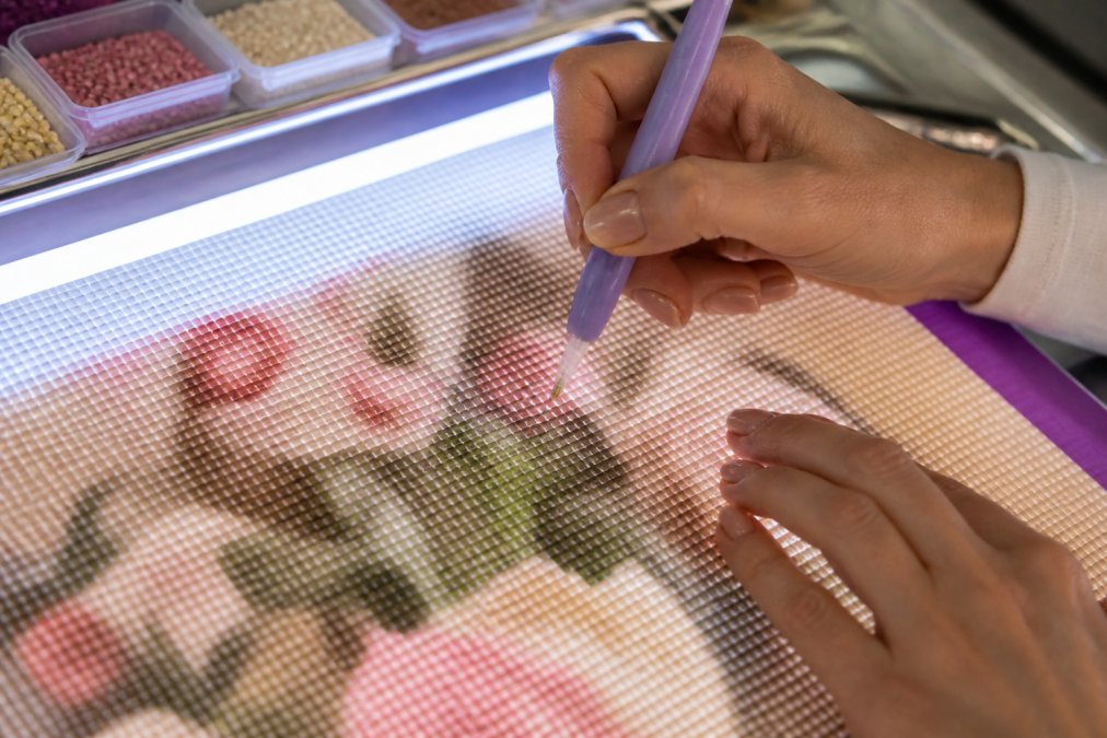 Close-up of hands using an ergonomic diamond painting pen with a light pad illuminating the canvas, showing proper grip and positioning over detailed drill placement