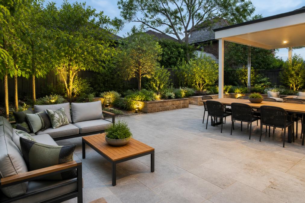 A residential backyard featuring a modern paved patio with comfortable outdoor seating arrangement, dining table, and lush landscaped garden borders with native plants creating defined entertaining zones