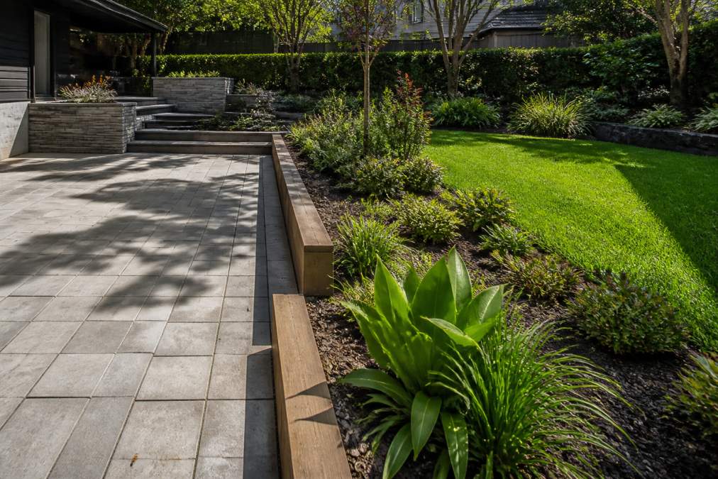 A residential garden displaying the contrast between hardscaped elements including stone paving, concrete retaining walls, and timber edging alongside softscaped areas with native plantings, lawn sections, and garden beds