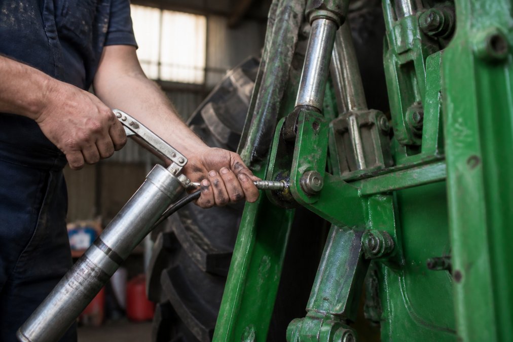 Mechanic's hands applying grease to tractor linkage pins and pivot points using an air-operated grease gun, with workshop equipment visible in the background