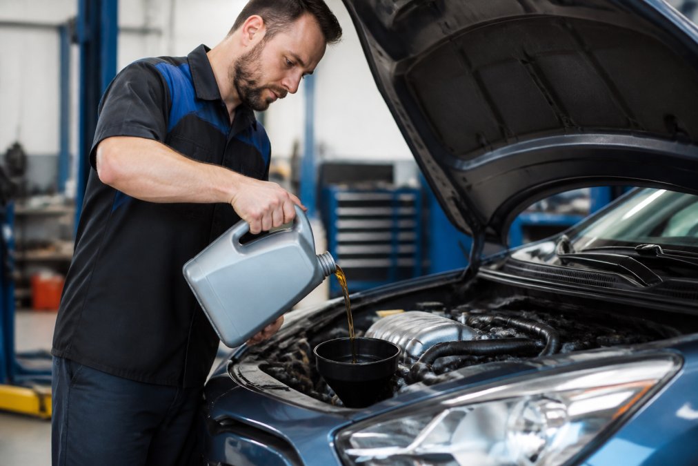 A mechanic in a professional garage pouring full synthetic motor oil from a jug into the engine bay of a Japanese sedan, with the hood open and a clean shop in the background
