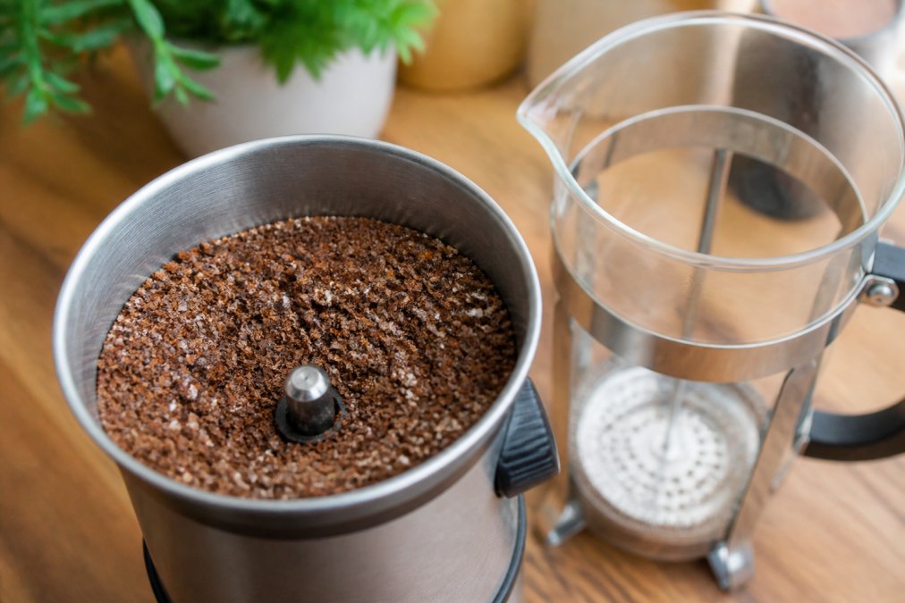 Close-up of coarse coffee grounds in a metal burr grinder hopper next to a glass French press, showing the granular texture and size of properly ground beans for immersion brewing