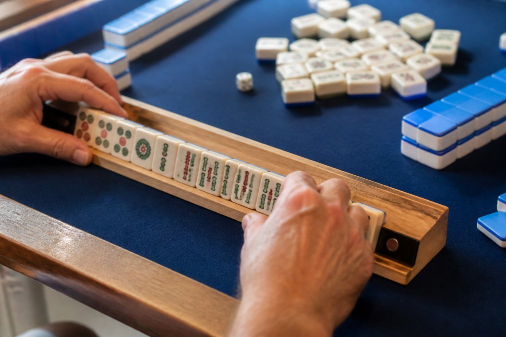 Close-up of hands arranging mahjong tiles on a wooden game rack with magnetic pushers during active gameplay, tiles sorted and visible on the rack