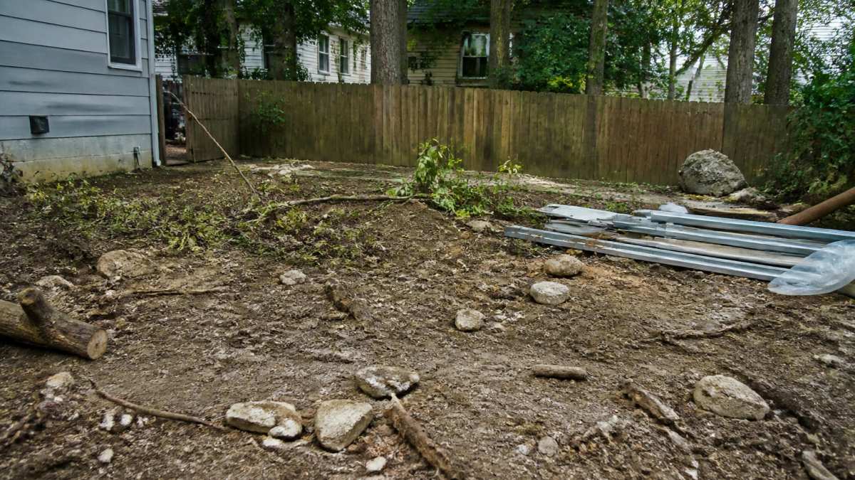 Residential yard with scattered debris, fallen branches, rocks, and construction materials across the ground before clearing and preparation
