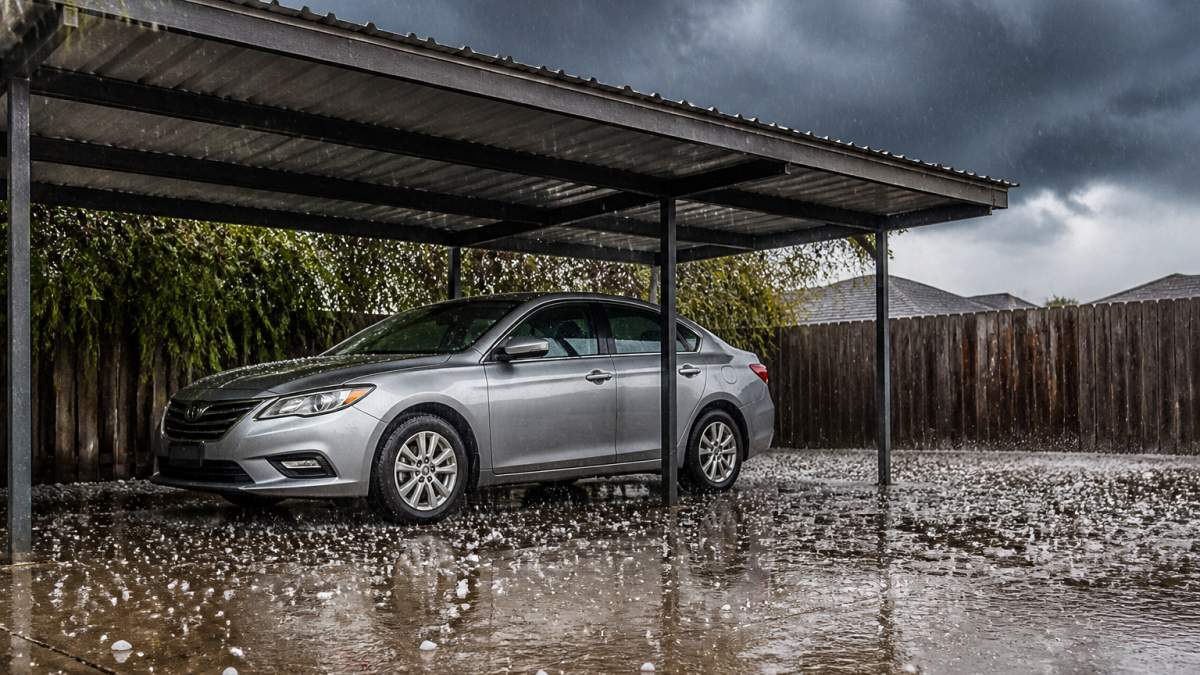 A silver sedan parked under a sturdy metal carport canopy while hailstones bounce off the concrete driveway, dark storm clouds overhead and wet pavement reflecting the gray sky