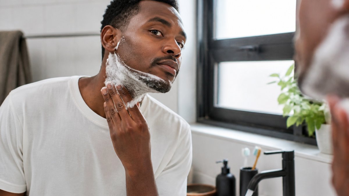Close-up of a Black man with coarse facial hair applying shaving cream to his jawline in a bright bathroom mirror, warm water running in the sink, focused and calm expression