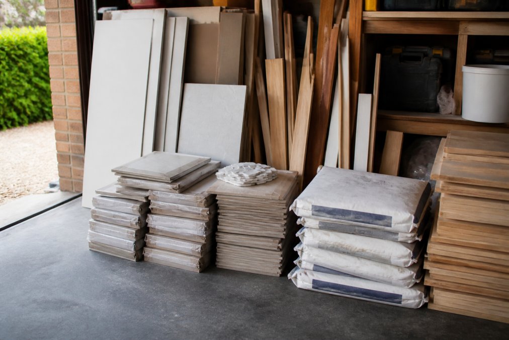 Neatly stacked leftover tiles, timber offcuts and unopened bags of grout arranged in a garage workshop, ready to be donated or sold on, with clear natural light from an open door