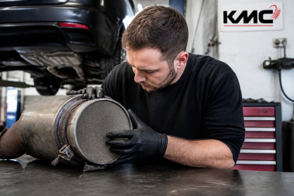 Close-up of a diesel particulate filter component being inspected by a technician during vehicle maintenance, with workshop tools and diagnostic equipment visible