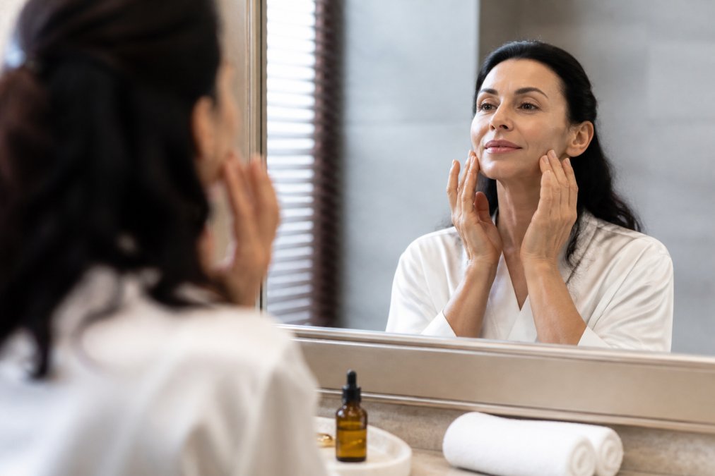 A woman in her forties applying facial massage with her fingertips in a softly lit bathroom, small bottle of facial oil on the counter, calm self-care moment