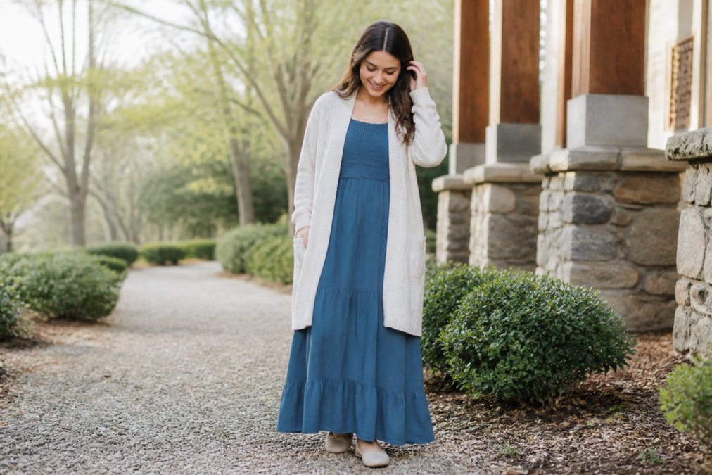 A woman wearing a long modest dress with a light cardigan layered over it, standing in a bright outdoor setting with soft natural light, relaxed and confident posture
