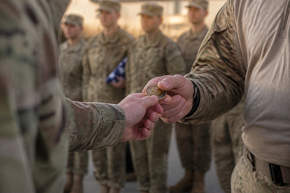 A close-up of a hand passing a custom military challenge coin to another service member during a ceremony, with the unit's insignia visible on the coin