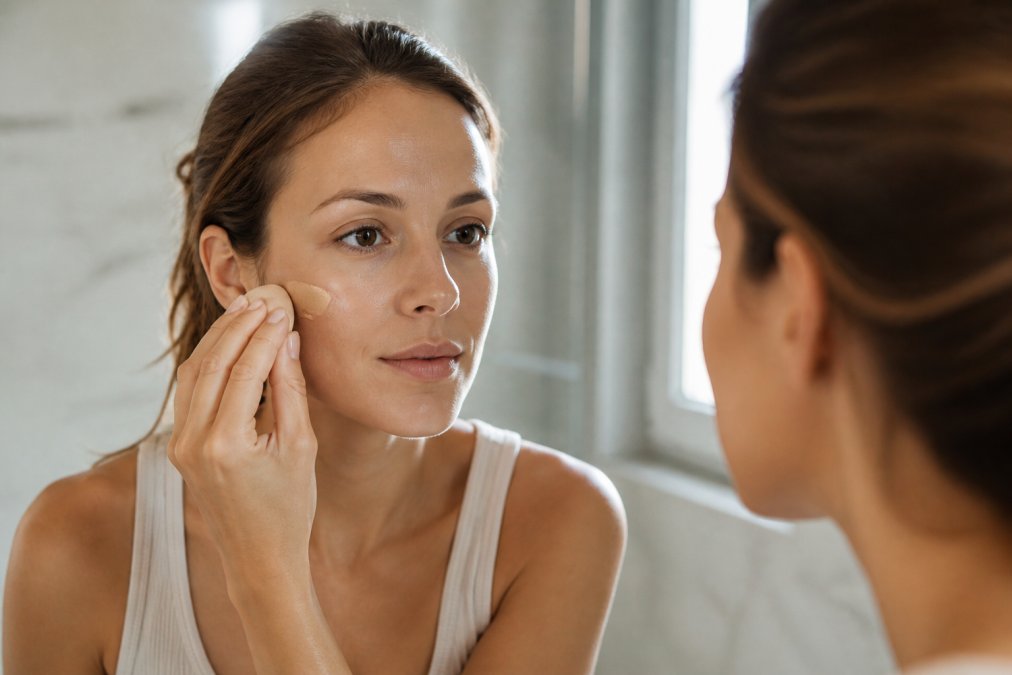 A close-up of a woman with glowing skin applying BB cream with a makeup sponge in a bright, modern bathroom