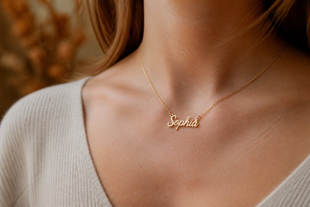 Close-up of a delicate custom name necklace displayed on a woman's neck, showing the personalized pendant and chain detail with soft natural lighting and warm background