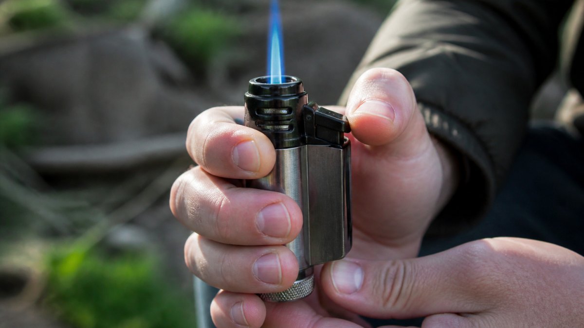 Close-up of a person's hands holding a jet torch lighter, examining the blue flame and metal body, with a blurred outdoor background