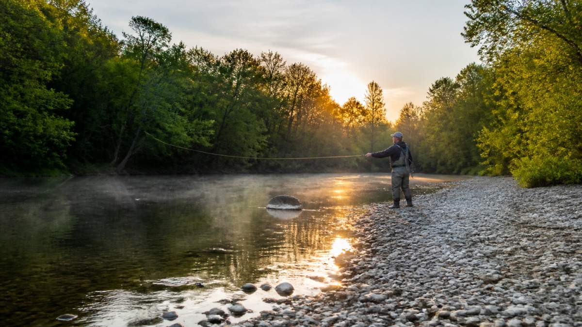 An angler fly casting at a scenic river fishing access point at sunrise