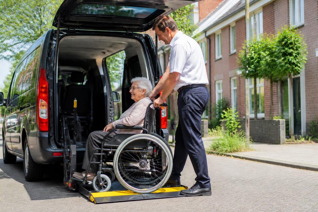 Een vriendelijke, professionele chauffeur in uniform helpt een oudere mevrouw in een rolstoel veilig de rolstoeltaxi in via een hydraulische lift, op een zonnige ochtend in een Utrechtse woonwijk