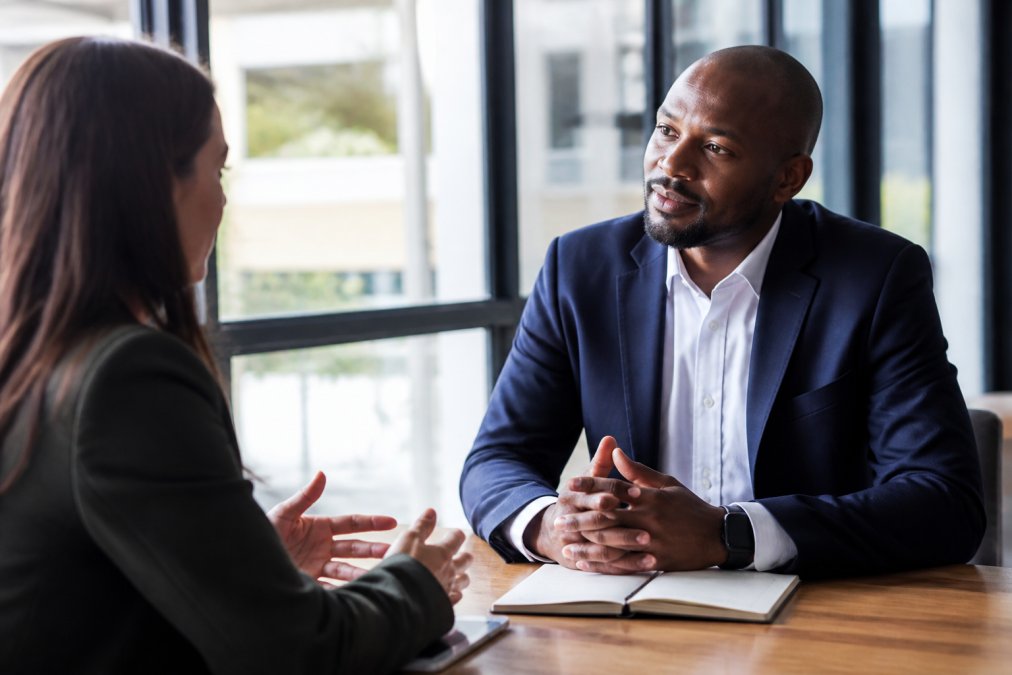 Business leader actively listening to team member during one-on-one meeting in modern office setting with natural sunlight, both seated at table with open body language