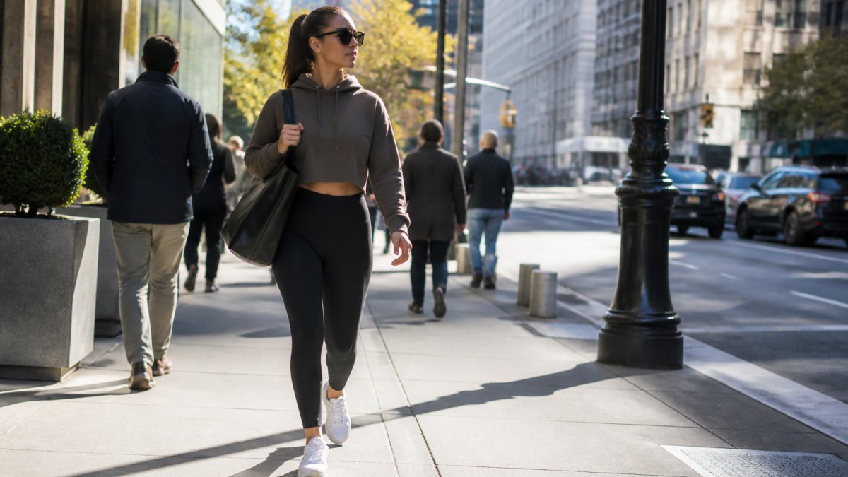 Confident person walking down a busy urban street wearing fitted leggings, a cropped hoodie, and clean white sneakers, carrying a structured bag, with natural morning light casting soft shadows on the pavement