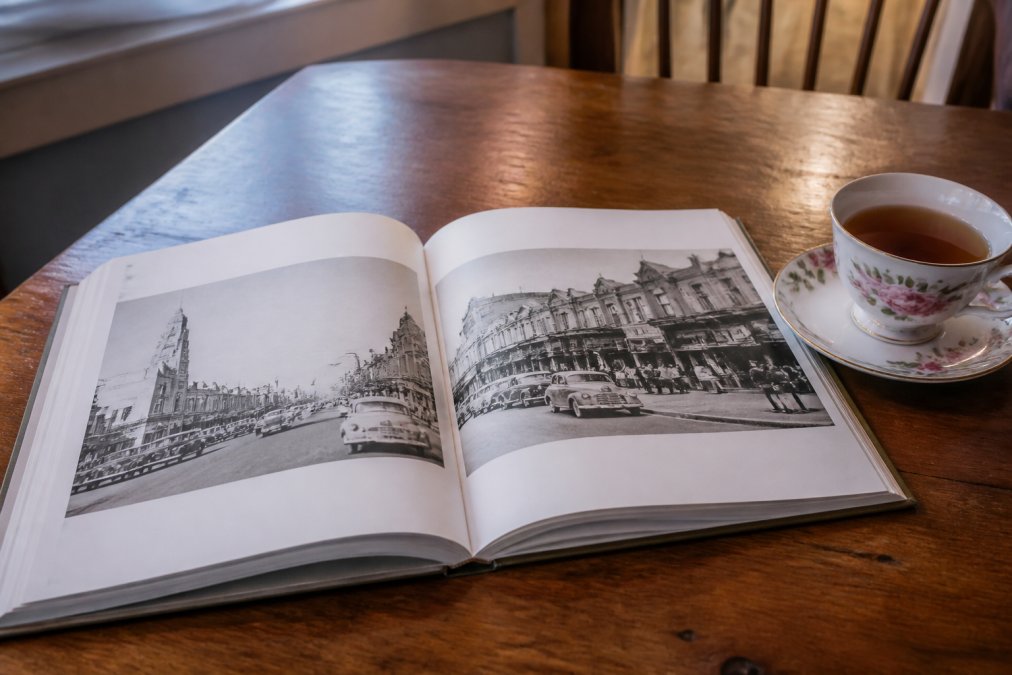 An open copy of a nostalgia photo book resting on a wooden table beside a cup of tea, with black and white photographs of 1950s Australian street scenes visible on the pages