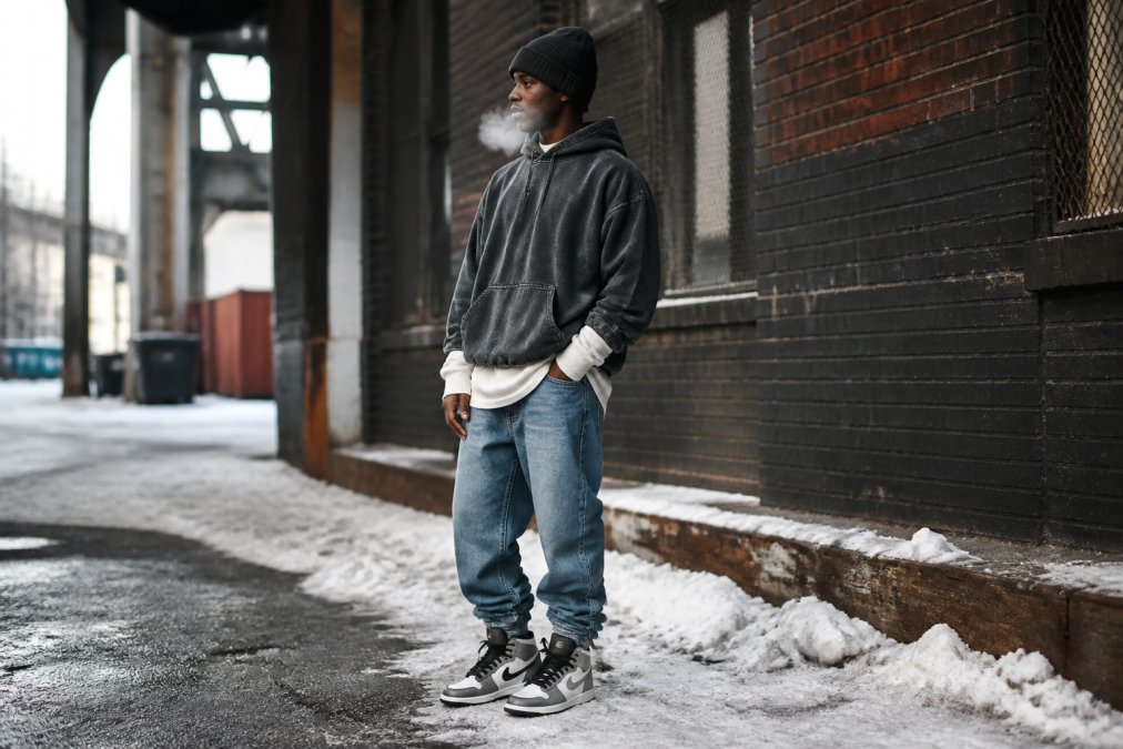 Person wearing layered winter streetwear outfit with oversized snow-washed hoodie over long-sleeve thermal, stacked denim, and high-top sneakers standing in urban cold-weather setting with visible breath