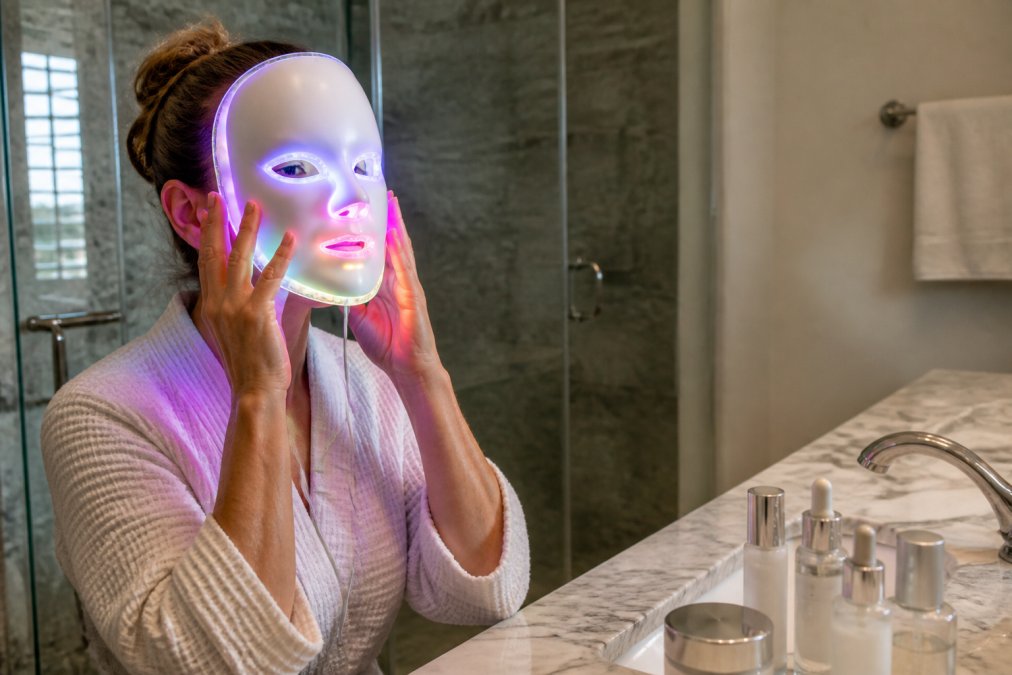 Woman in her 40s applying a 7-color LED facial mask at home in a serene bathroom setting, with soft lighting and skincare products on marble counter