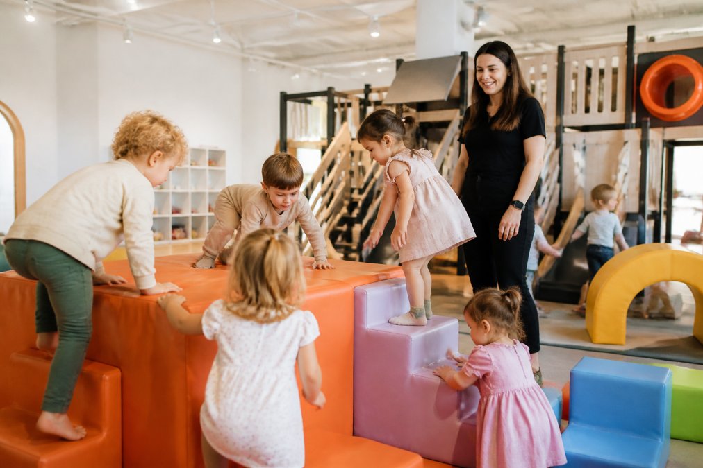 Young children playing safely in a bright, climate-controlled indoor play area with soft play structures, foam blocks, and climbing equipment while a teacher supervises nearby