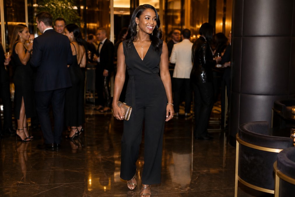 Woman wearing a sleek black jumpsuit styled with strappy heels, statement jewelry, and a metallic clutch at an upscale venue with ambient lighting and elegant décor