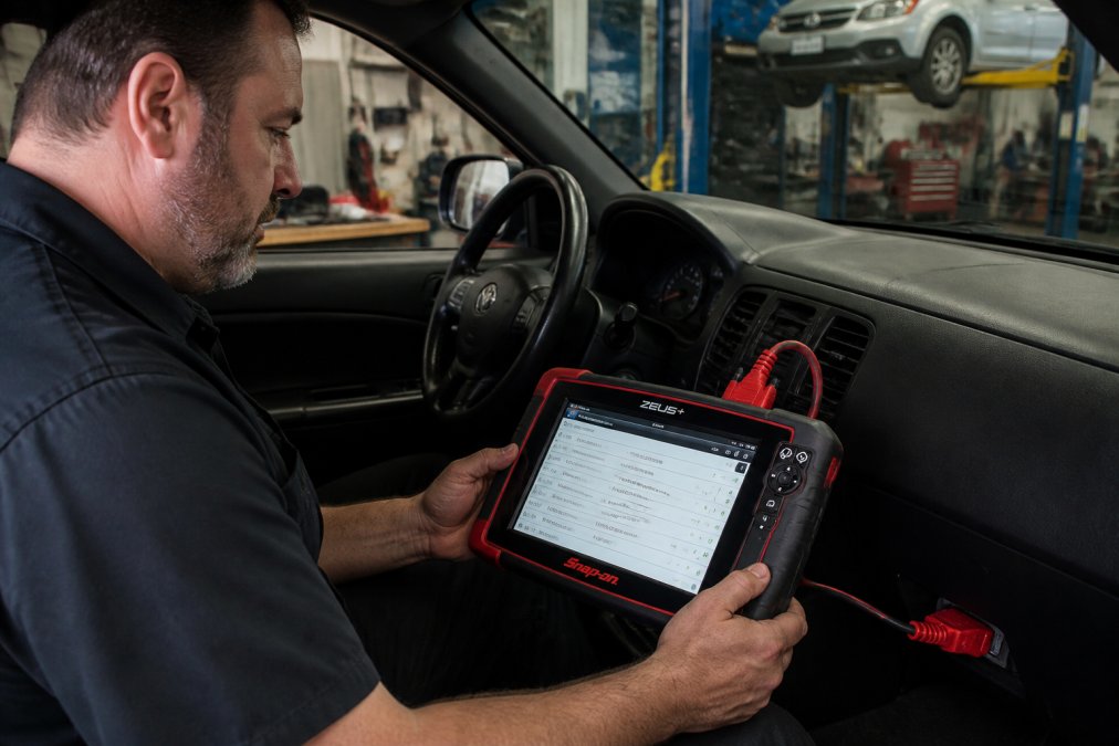 Technician using Snap-on ZEUS+ diagnostic scanner connected to vehicle's data link connector, tablet screen displaying fault codes in collision repair shop with diagnostic equipment visible in background