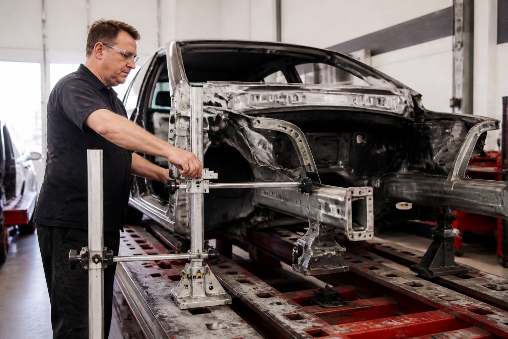 Auto body technician operating Spanesi Touch 3D electronic measuring system on damaged vehicle frame, measurement probes positioned on chassis reference points with real-time measurement data visible on monitor in collision repair shop