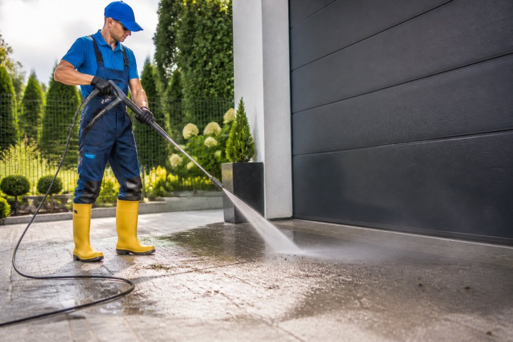 A uniformed pressure cleaning technician operating industrial-grade high-pressure equipment on a residential driveway, water and displaced grime spraying across grey concrete in bright afternoon sunlight