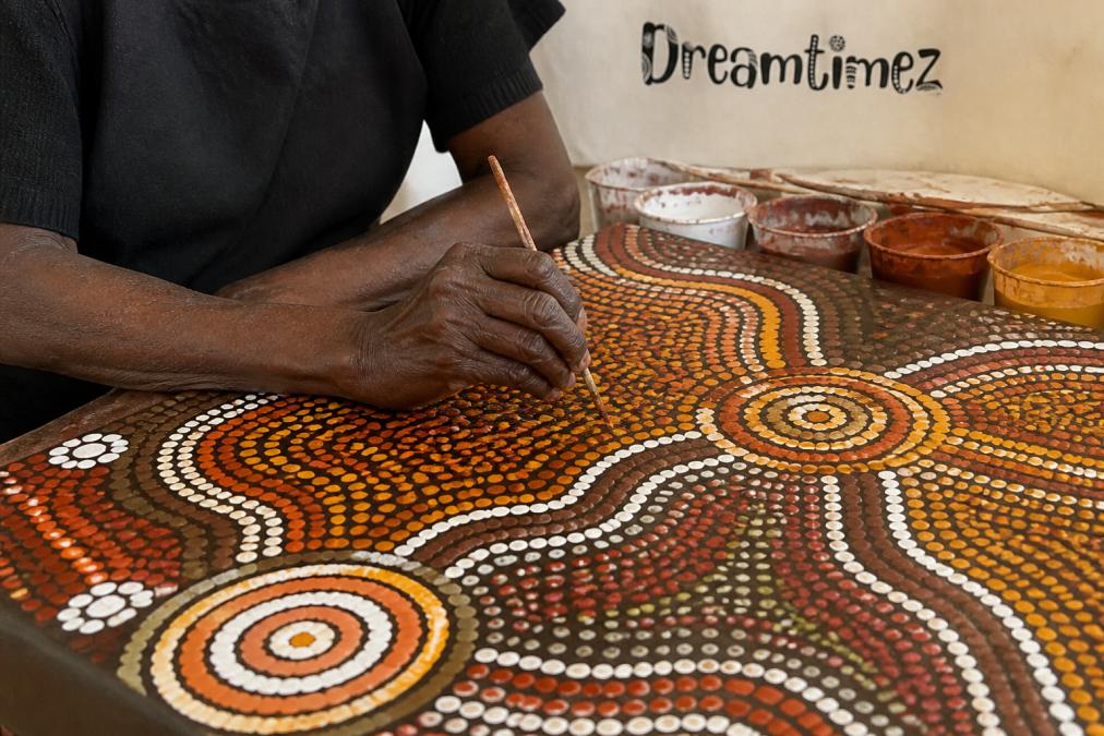 Close-up of authentic Aboriginal artwork displaying intricate dot painting patterns with natural ochre pigments on canvas, hands of artist visible applying traditional techniques
