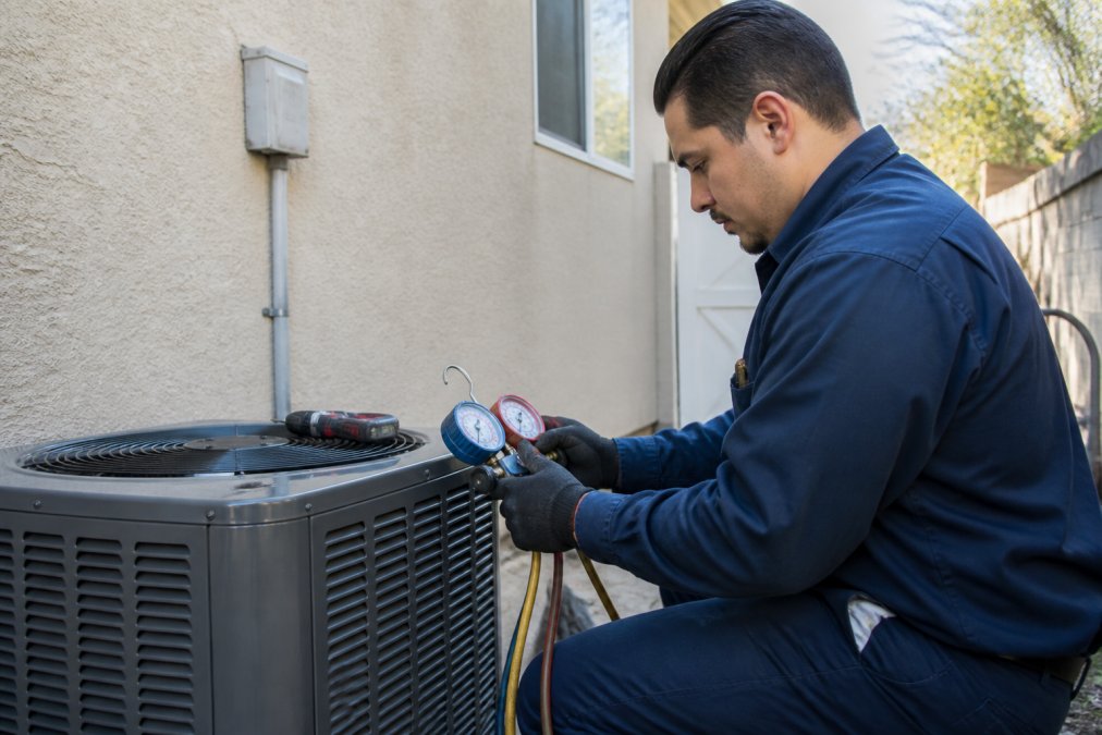 Licensed HVAC technician in uniform performing diagnostic inspection on outdoor AC condenser unit at residential home, checking refrigerant levels with gauges