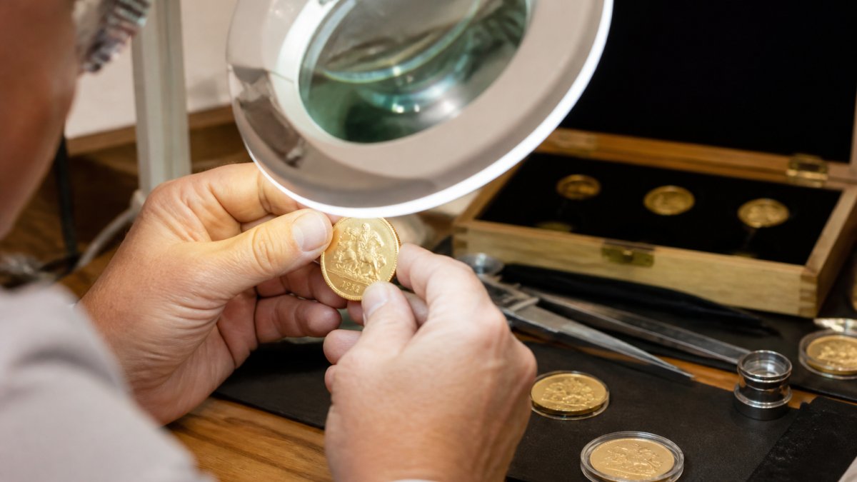 Professional jeweller's hands holding a gold bullion coin under a magnifying lamp, examining the coin's surface markings and edge detail in a well-lit workshop setting