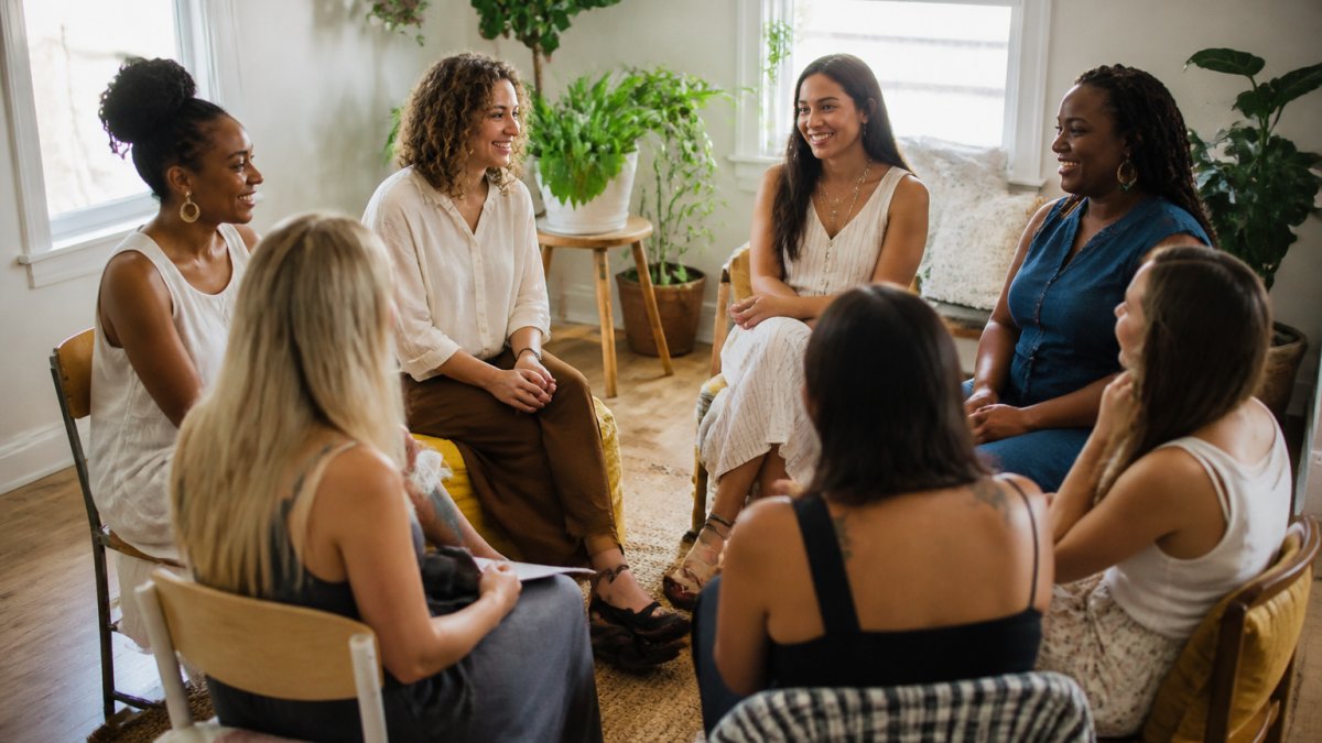 Diverse group of women in a circle having a supportive conversation in a warm, welcoming indoor setting with natural light, genuine smiles and engaged body language
