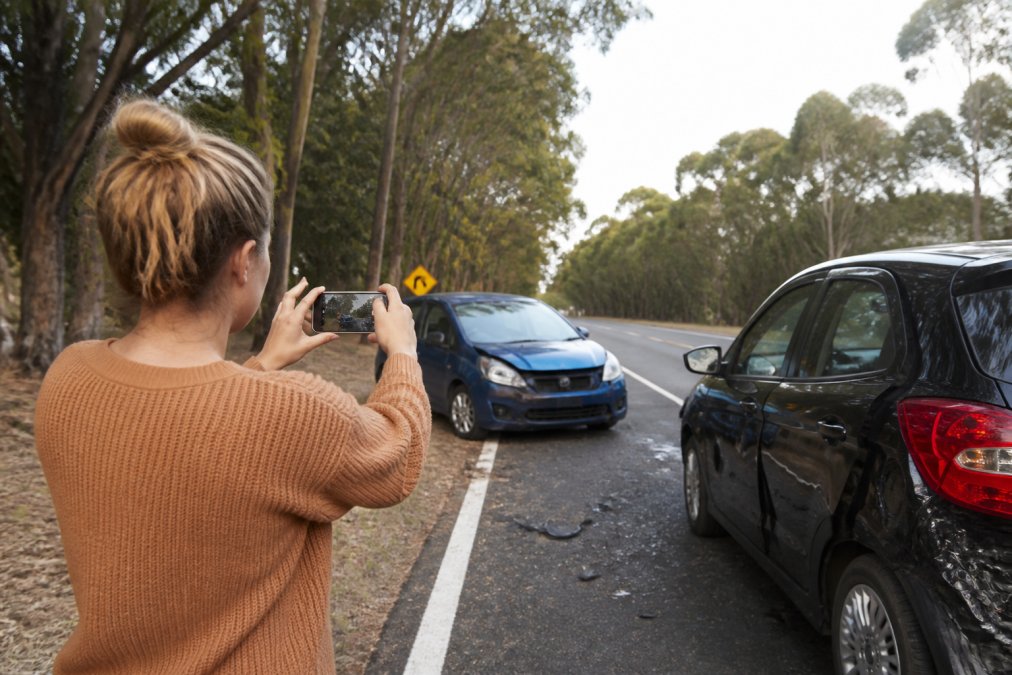 A driver standing safely outside a damaged vehicle on a regional road, taking photos of the accident scene with their phone while another vehicle sits damaged nearby