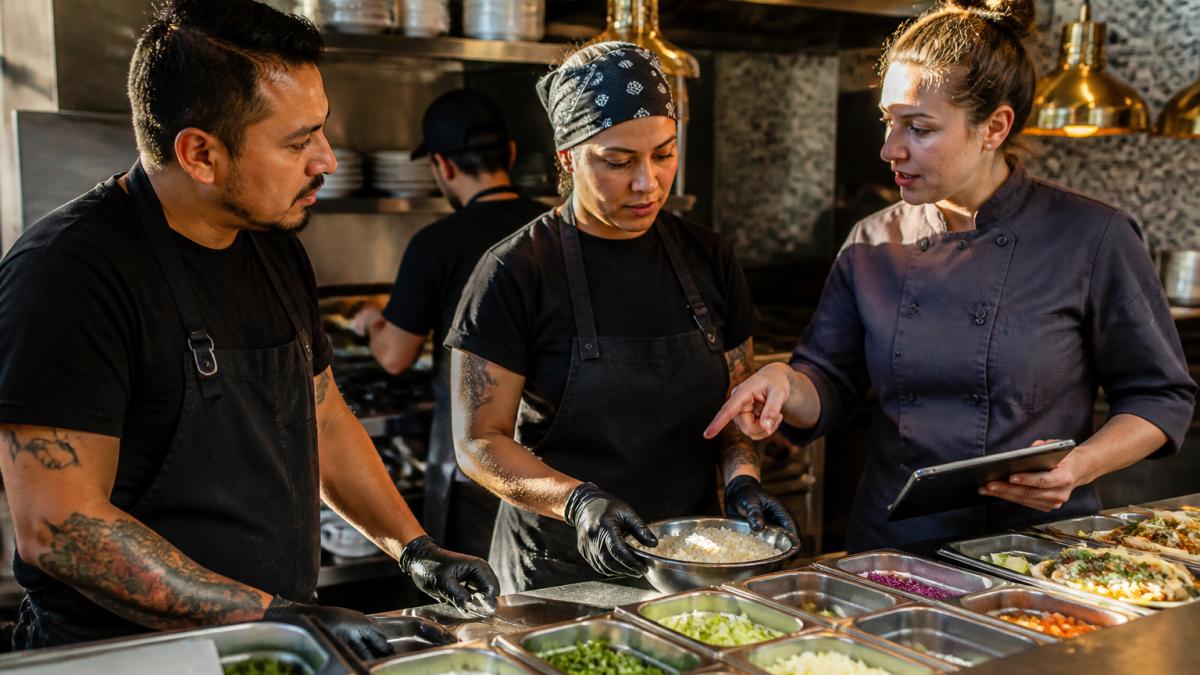 Professional kitchen staff collaborating during food preparation, wearing aprons and discussing meal components at a prep station with organized ingredients visible
