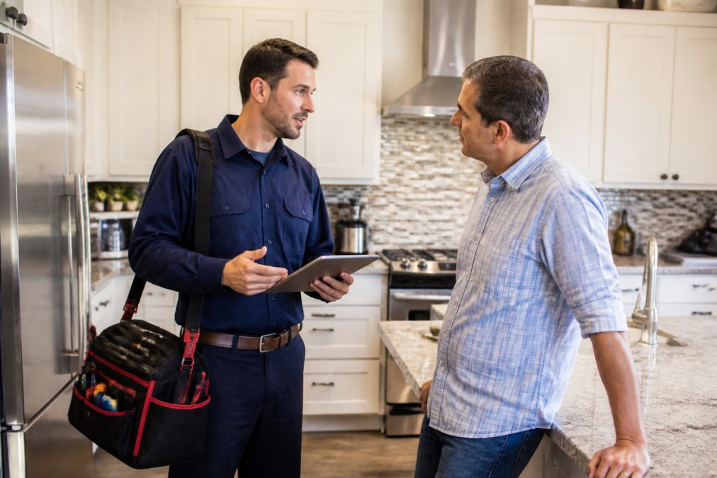Uniformed service technician with tablet and tool bag standing in residential kitchen, discussing project with homeowner, showing professional appearance and digital-first approach