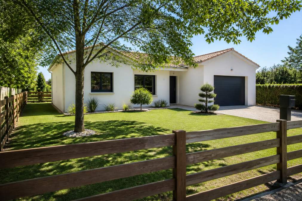 Façade d'une maison plain-pied moderne avec garage intégré à droite, jardin clôturé avec clôture en bois bien entretenue, pelouse verte, jour ensoleillé avec ombre des arbres