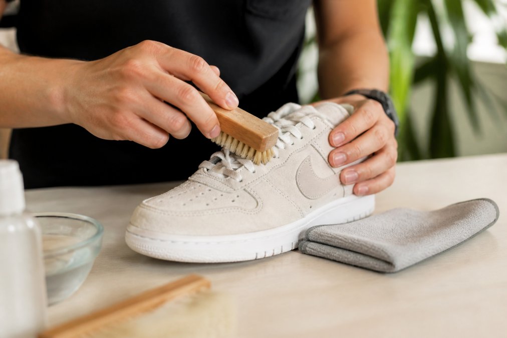 Gros plan de mains nettoyant délicatement une sneaker Nike en daim blanc avec une brosse douce et un chiffon microfibre, posée sur un bureau épuré avec un éclairage doux et naturel