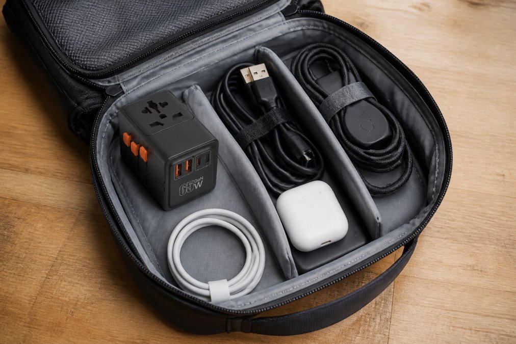 Vue de dessus d'un sac de voyage ouvert sur une surface en bois, avec un petit adaptateur universel, des câbles enroulés et des accessoires tech soigneusement organisés à l'intérieur