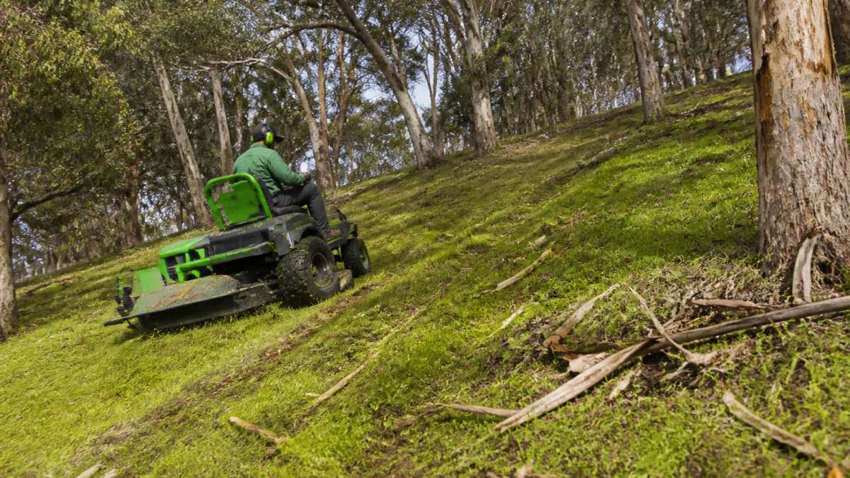 A ride-on mower with a slasher attachment working across a steep, sloping acreage block with scattered gum trees and fallen bark, the operator navigating around obstacles on a rural property