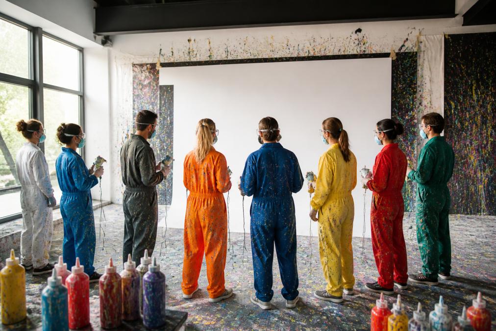 Gruppe von Teilnehmern in bunten Kitteln mit Schutzausrüstung, bereit zur Arbeit mit Farbschleudern, großformatige weiße Leinwand im Hintergrund, natürliche Beleuchtung in modernem Kunstraum