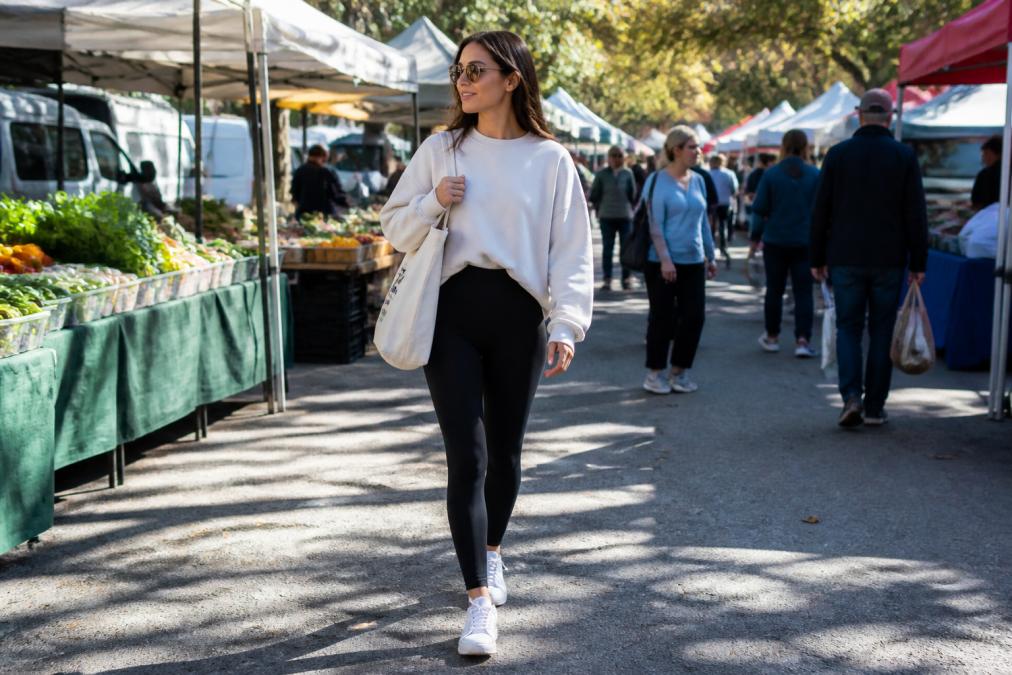 A woman wearing high-waisted black matte leggings, an oversized cream sweatshirt, and white low-top sneakers, carrying a canvas tote bag while walking through a sunlit weekend outdoor market with vendors in the background