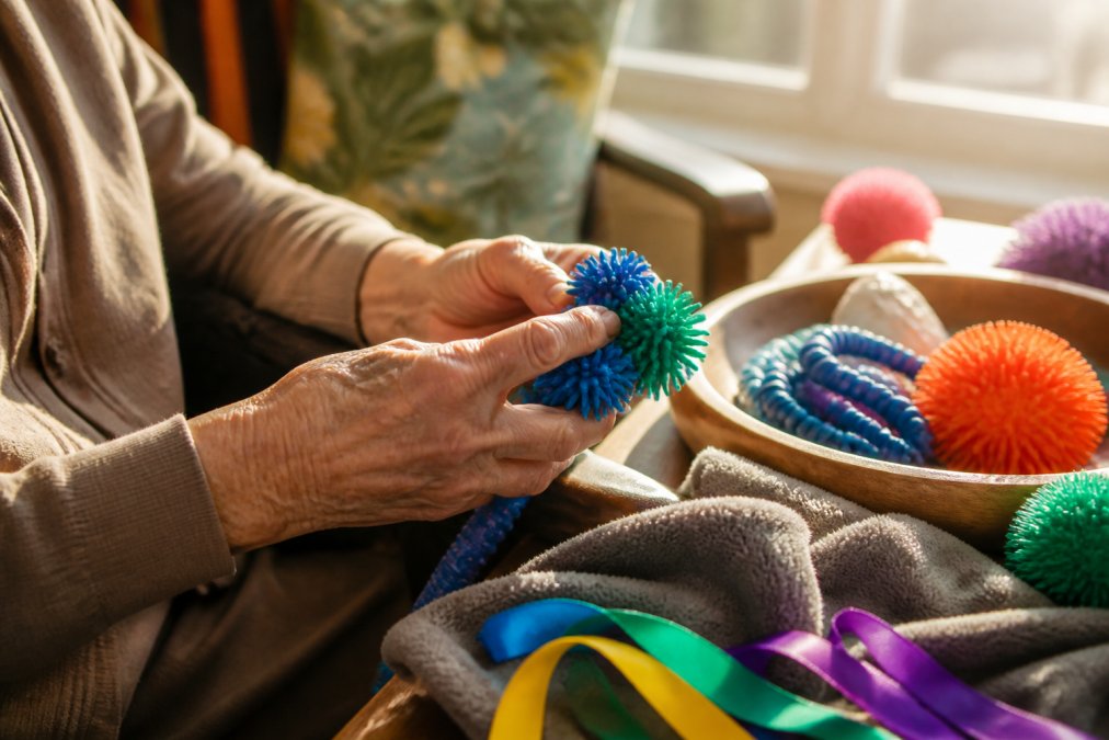 Close-up of an older adult's weathered hands gently holding and exploring colorful textured fidget items, soft fleece, smooth beads, silky ribbons, in natural afternoon sunlight from a window