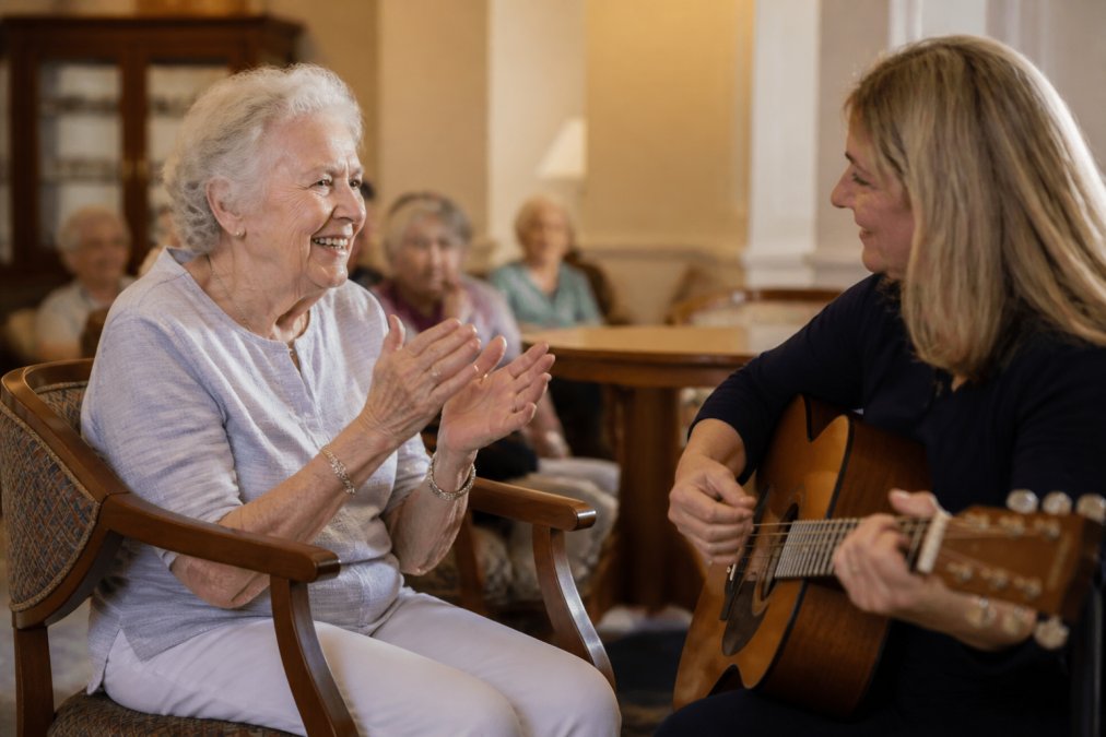 An elderly woman smiling and clapping her hands while a caregiver plays guitar beside her in a warmly lit common room, warm afternoon light casting a soft glow over both figures