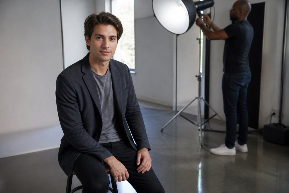 Actor in professional wardrobe sitting confidently in well-lit studio, making direct eye contact with natural, relaxed expression while photographer adjusts lighting in background