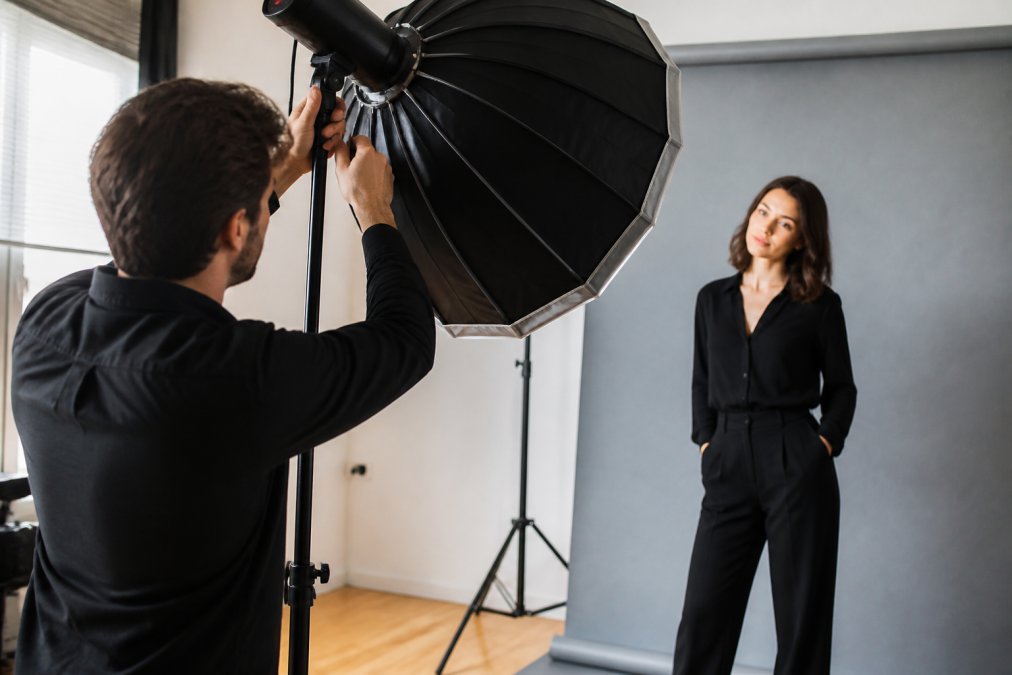 Professional photographer adjusting studio lighting equipment while actor poses confidently in background, demonstrating technical setup and direction process in modern studio