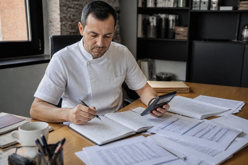 Gerente de restaurante en su oficina revisando una laptop con hojas de cálculo de costos de ingredientes esparcidas sobre el escritorio, sosteniendo una calculadora, con notas manuscritas de márgenes y precios